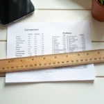 Règle en bois vintage montrant des graduations en centimètres et en pouces sur un bureau blanc, avec une table de conversion imprimée